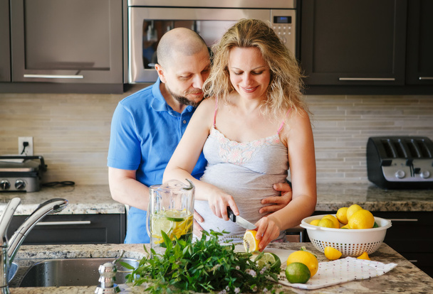 Portrait of smiling white Caucasian couple two people pregnant woman with husband cooking food fruit lemon juice, standing in kitchen, lifestyle healthy pregnancy happy life concept - Photo, Image