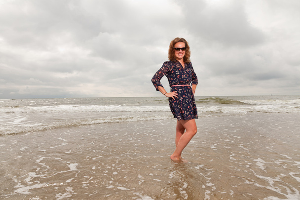Pretty young woman enjoying outdoor nature near the beach. Standing in the water. Red hair. Wearing dark blue dress and black sunglasses. Cloudy sky. - Фото, изображение