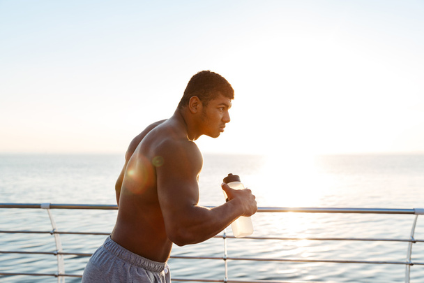 Shirtless african sportsman with bottle of water running on pier - Foto, Bild