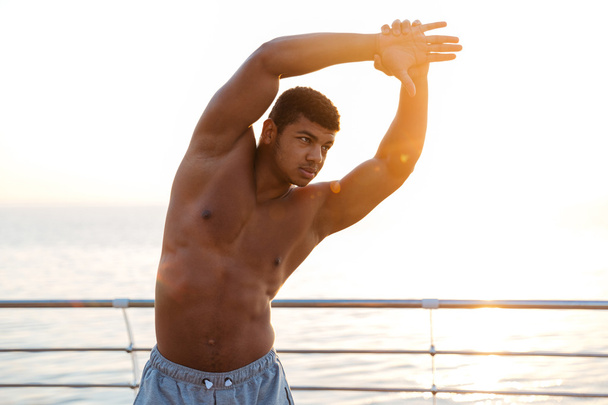 Handsome muscular african man athlete doing stretching exercises on pier - Φωτογραφία, εικόνα