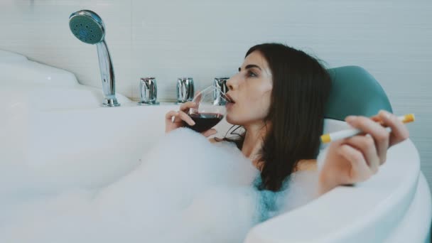 Girl taking bath full of foam. Smoke electronic cigarette, drink red wine. - Imágenes, Vídeo