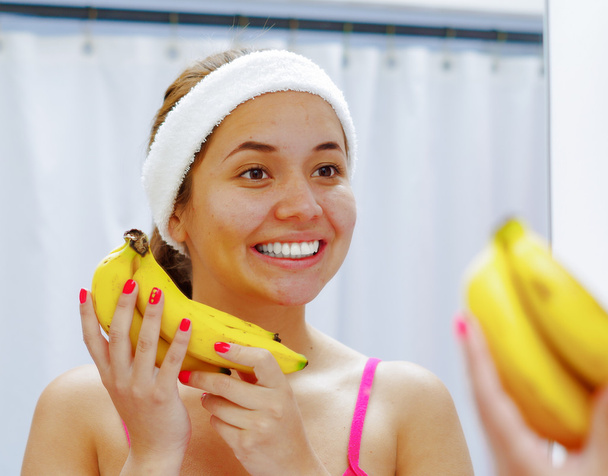 Attractive young woman wearing pink top and white headband, holding bananas, looking in mirror smiling - Foto, Imagem