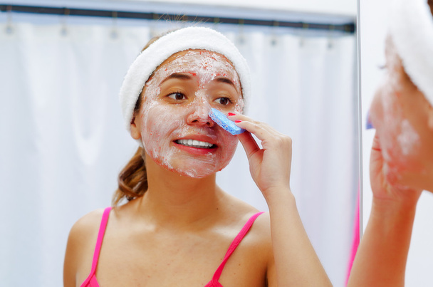 Attractive young woman wearing pink top and white headband removing cream from face using sponge, looking in mirror smiling - Foto, afbeelding