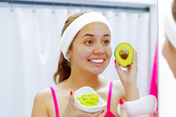 Attractive young woman wearing pink top and white headband, holding up avocado used for skin treatment, looking in mirror smiling - Photo, image