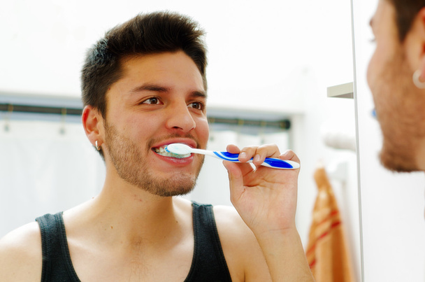 Handsome young man wearing black singlet top looking in mirror smiling while brushing teeth - Fotografie, Obrázek