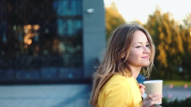Young beautiful girl in a yellow jacket near office staying and smiling at the balkony with the coffee. Smiling business woman with cup of tea at the business center. Bright sunlight in the morning. - Filmmaterial, Video