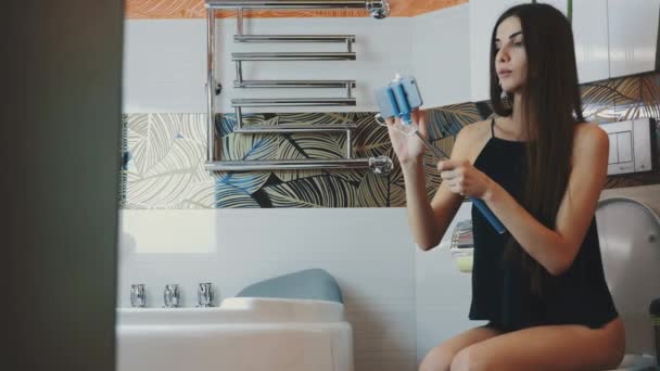 Attractive girl sitting on toilet in bathroom with blue monopod for selfie - Кадри, відео