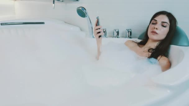 Brunette girl enjoy bath full of foam. Tap in smartphone. Relax. Resting. - Footage, Video