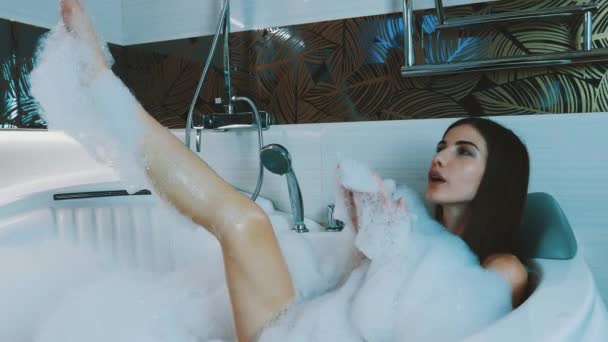 Brunette young girl taking bath full of foam in bathroom. Blow bubbles from foam - Footage, Video