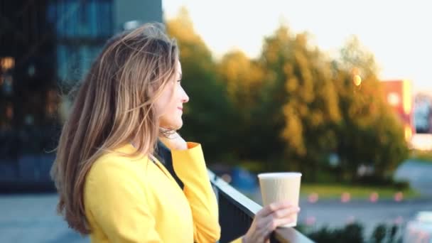 Slow motion. Young beautiful girl in a yellow jacket near office staying and smiling at the balkony with the coffee. Smiling business woman with cup of tea at the business center. - Video, Çekim