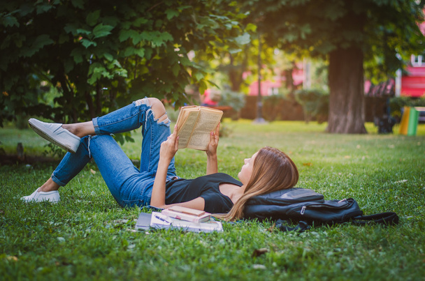 студентка в парке, лежащая на траве и читающая книгу, лежащая рядом со своим рюкзаком и стопкой тетрадей. Концепция образования
 - Фото, изображение