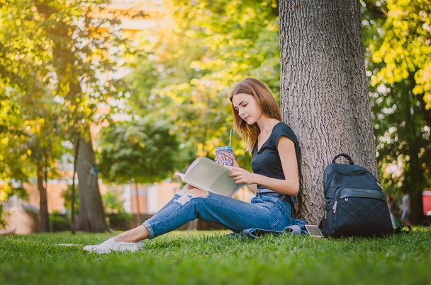 Boldog lány diák ül a füvön, közel a fához a parkban és olvas. Oktatási koncepció - Fotó, kép