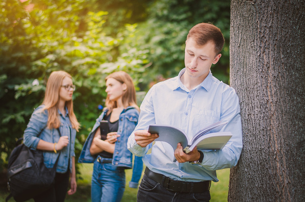 счастливый улыбающийся мальчик студент в брекетах, рубашке и брюках с книгой в летнем парке, за ним стоят девочки и общаются
 - Фото, изображение