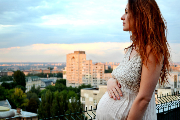 Joven chica pelirroja embarazada hermosa y feliz sobre la vista de la puesta del sol y la ciudad mirando hacia adelante de la maternidad futura y esperando a su bebé en un vestido blanco romántico
   - Foto, Imagen