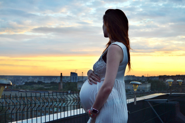 Mladá, krásná a šťastná těhotné redhaired dívka za výhled na západ slunce a město hledá vpřed budoucí mateřství a očekává její dítě v romantické bílé šaty   - Fotografie, Obrázek