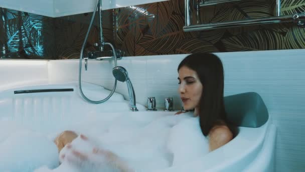Happy girl taking bath full of foam in bathroom. Washing legs. Blow soap foam. - Footage, Video