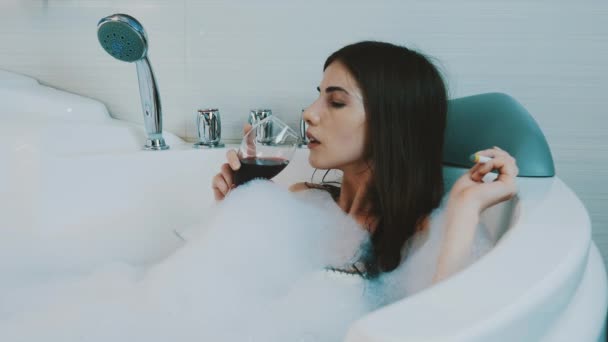 Girl drink red wine in bath full of foam. Smoking electronic cigarette. Bathroom - Πλάνα, βίντεο