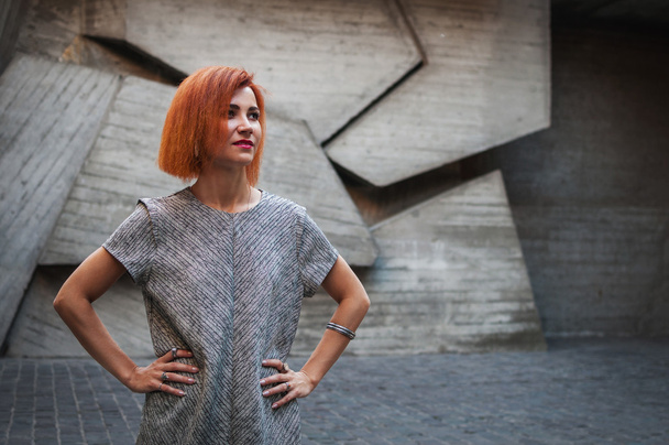 beautiful redhead girl wearing in a gray dress posing against background of concrete wall - Photo, Image