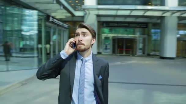 Jóképű üzletember beszél a modern üzleti központjához közel, a szúró telefon felhőkarcoló. A fényképezőgép mozgó körül az ember (Steadicamnél lövés) - Felvétel, videó