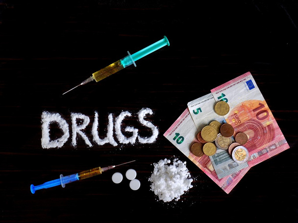 Cocaine drug powder in shaped drug word and pile of this cocaine between two injections, pills and euro money, banknotes and coins on black background - Photo, Image