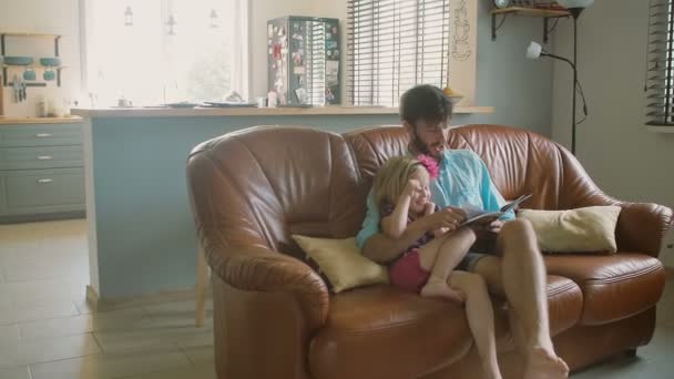 A young handsome father is sitting on the sofa reading a book to his precious little daughter. Slow mo, Steadicam shot - Video