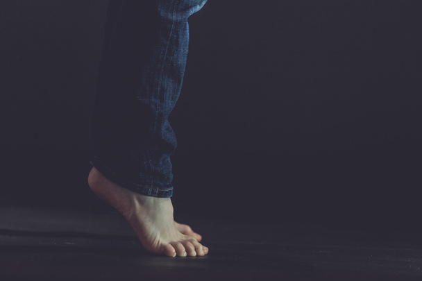 Κοντινό πλάνο γυμνά πόδια καθώς στέκεται όρθιος στο πάτωμα σκούρο - Φωτογραφία, εικόνα