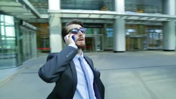 Handsome businessman in sunglasses is talking on the phone near the modern business center, skyscraper. The camera is moving around man (steadicam shot) - Video