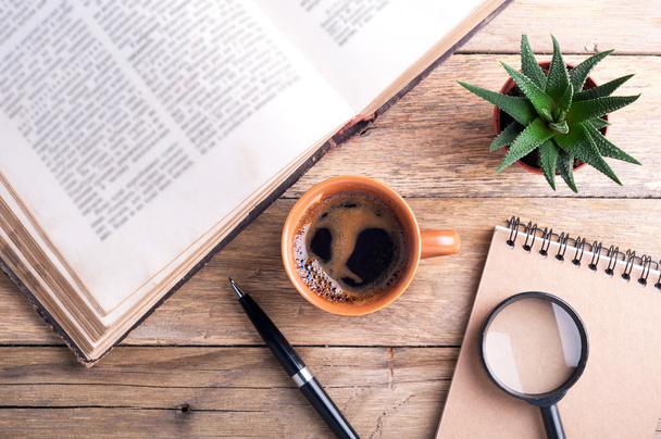 Cup of coffee, old open book, succulent, pen, notebook and magnifier on rustic wooden background. Office desk table - Photo, Image
