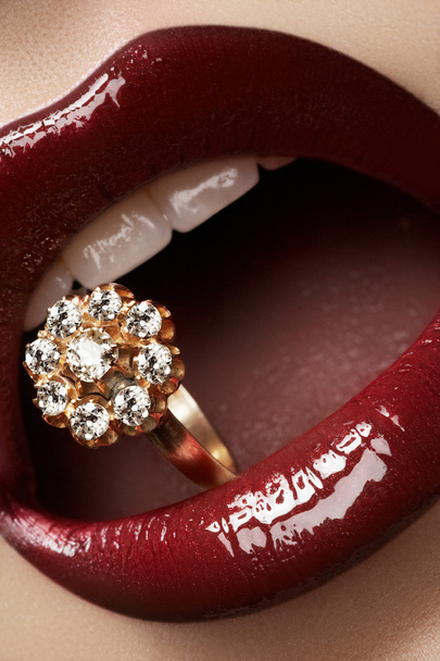 Close-up of beautiful woman's lips with bright fashion dark red glossy makeup. Macro lipgloss cherry make-up. Mouth with wedding gold diamond ring. Jewelry accessories - 写真・画像