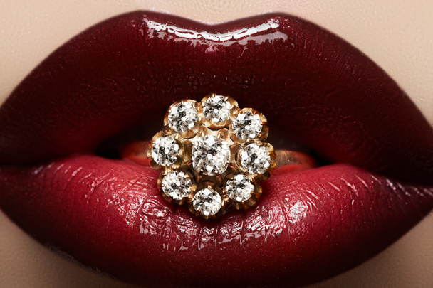 Close-up van mooie vrouw lippen met heldere mode donker rode glanzende make-up. Macro lipgloss cherry make-up. Mond met bruiloft gold diamond ring - Foto, afbeelding