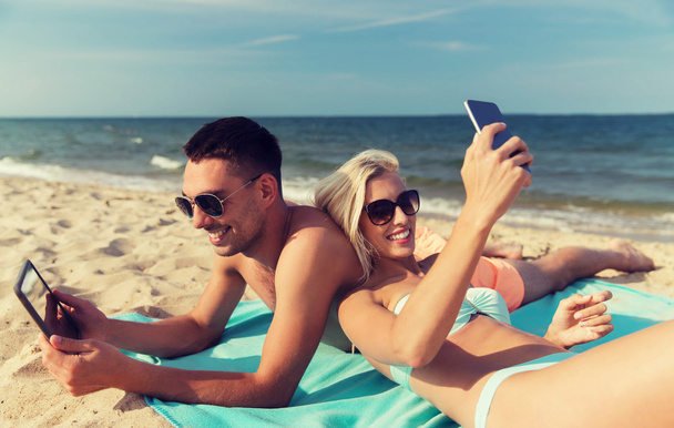 happy couple with modern gadgets lying on beach - 写真・画像