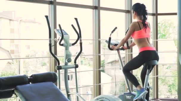Sexy girl doing exercise with exercise bike in the gym. Look behind her - Кадры, видео
