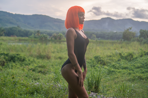 Sexy beautiful woman in modern futuristic style posing on green field over amazing sunset sky background. Creative look of tattooed woman wearing black bodysuit and orange wig - Photo, Image