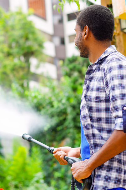 Man wearing square pattern blue and white shirt holding high pressure water gun, pointing liquid to green garden - Foto, Imagem