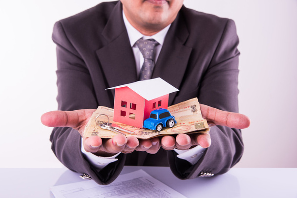 home and car buying concept, indian man holding indian currency in both hands and model house and toy car over it, closeup and selective focus - Photo, Image