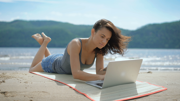 Μια όμορφη σέξι νεαρή κοπέλα στοχαστικό είναι laying επί της tommy σε μια παραλία που χρησιμοποιούν φορητό υπολογιστή - Πλάνα, βίντεο