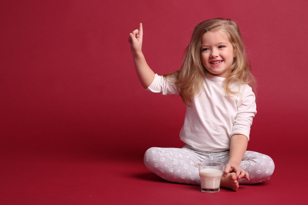 Baby girl in pajamas with glass of milk showing finger. Red background - Photo, Image