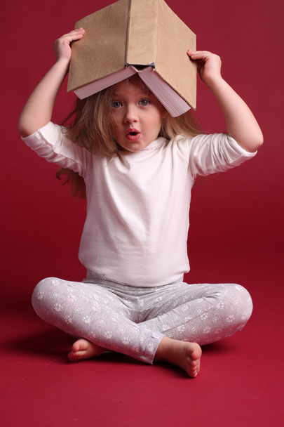 Kid in sleepwear with a book on her head. Red background - Foto, afbeelding