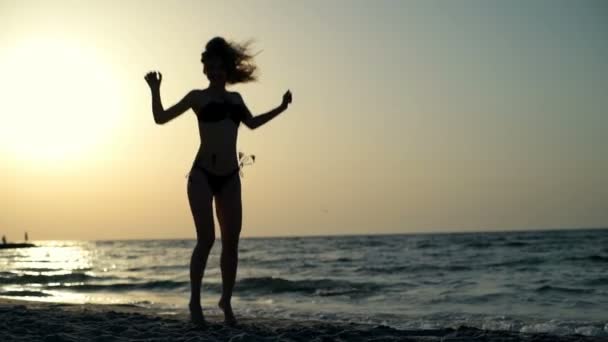 Sziluettjét a fiatal gyönyörű lány fürdőruha jumping, séta a tengerparton - Felvétel, videó