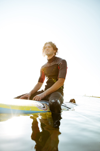 Handsome young surfer man in swimwear sitting on surf board - Zdjęcie, obraz