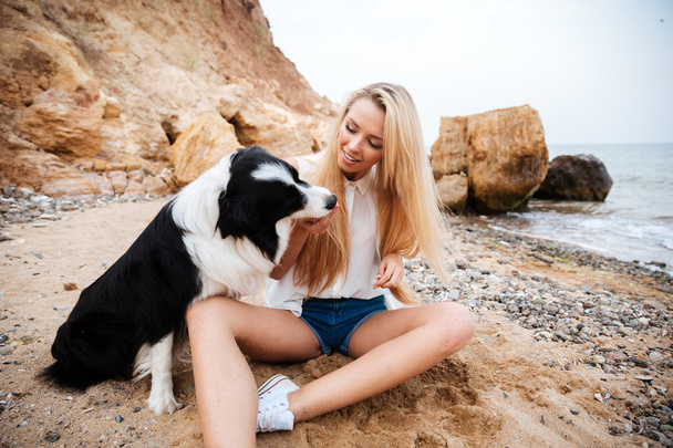 Smiling woman sitting and playing with dog on beach - Fotoğraf, Görsel