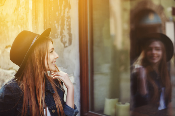 Portrait of young smiling woman looking at the shop window. Model wearing stylish wide-brimmed black hat. City lifestyle. Close up. Toned - Photo, Image