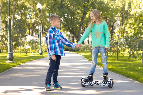 Chica y niño caminando de la mano. Una chica está parada en un scooter giratorio. Se sonríen.
 .  - Foto, imagen