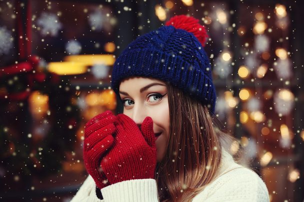 Street portrait of young beautiful woman warming hands with her mouth. Model looking at camera. Lady wearing stylish classic winter knitted clothes. Christmas concept. Snowfall. Close up - Photo, Image