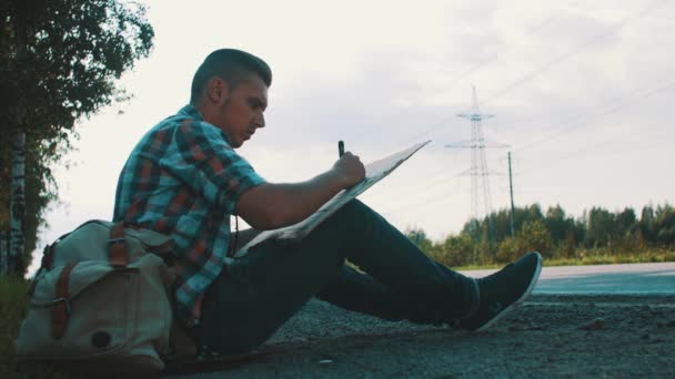 Man sit at road in countryside write on cardboard plate. Hitchhiking. Backpack. - Séquence, vidéo