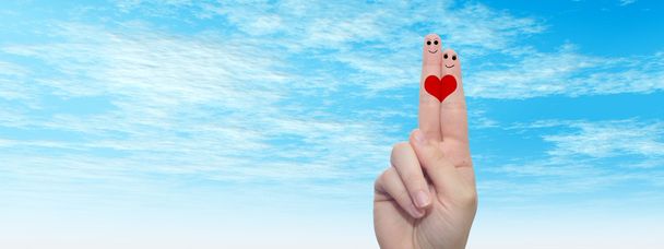 Έννοια ή εννοιολογική ανθρώπινη ή γυναικεία χέρια με δύο δάχτυλα βαμμένα με κόκκινη καρδιά και χαμογελαστά πρόσωπα πάνω από το σύννεφο μπλε ουρανό φόντο πανό - Φωτογραφία, εικόνα