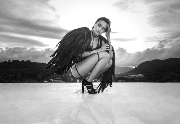 Black and white photo of beautiful seductive angel woman wearing lingerie and leather belts crouching on the roof over cloudy sky - Photo, Image