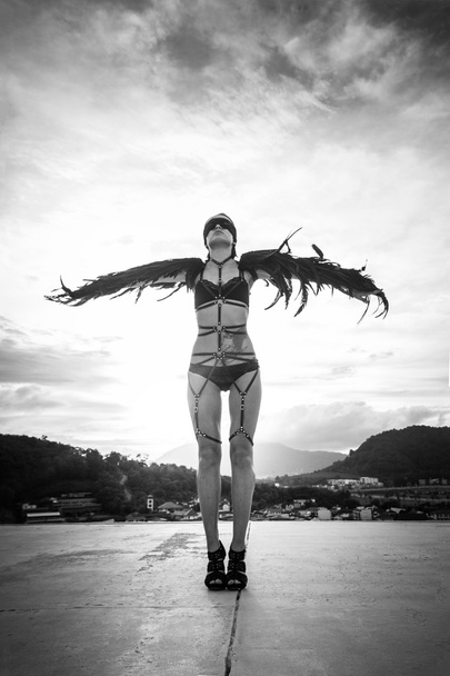 Black and white photo of beautiful seductive angel woman wearing lingerie and leather belts standing on the roof with wind in her wings over cloudy sky - Foto, Bild