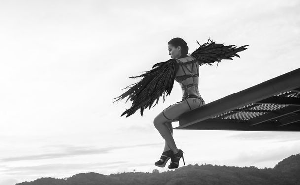 Black and white photo of beautiful seductive angel woman wearing lingerie and leather belts sitting on the roof edge with wind in her wings over cloudy sky - Photo, Image