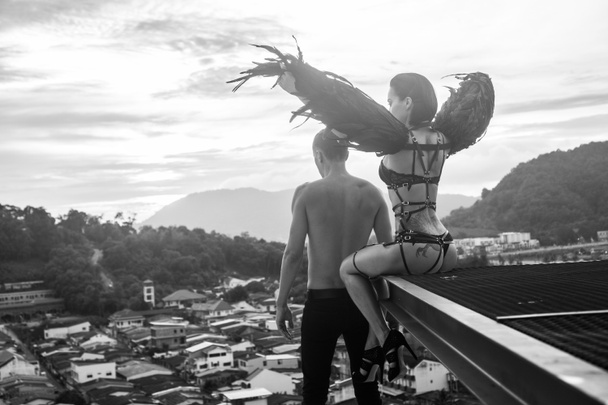 Black and white photo of romantic scene with shirtless man and sensual angel woman wearing lingerie, leather belts and high heels on the rooftop over sky and city landscape background - Photo, Image